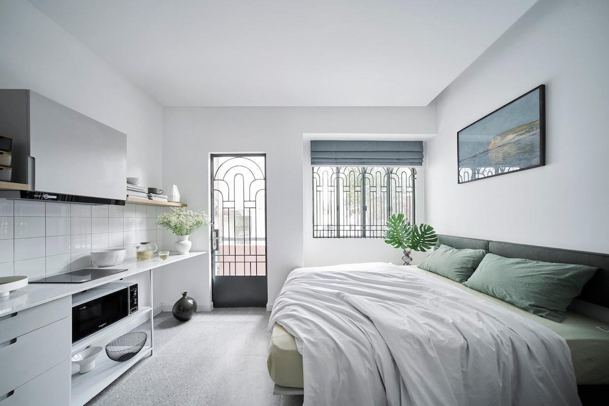 A minimalistic bedroom features a spacious bed dressed in light green linens. Natural light filters through a window with a decorative grille, illuminating the sleek cabinetry and a small kitchenette area. A plant adds a touch of greenery, enhancing the calm ambiance.