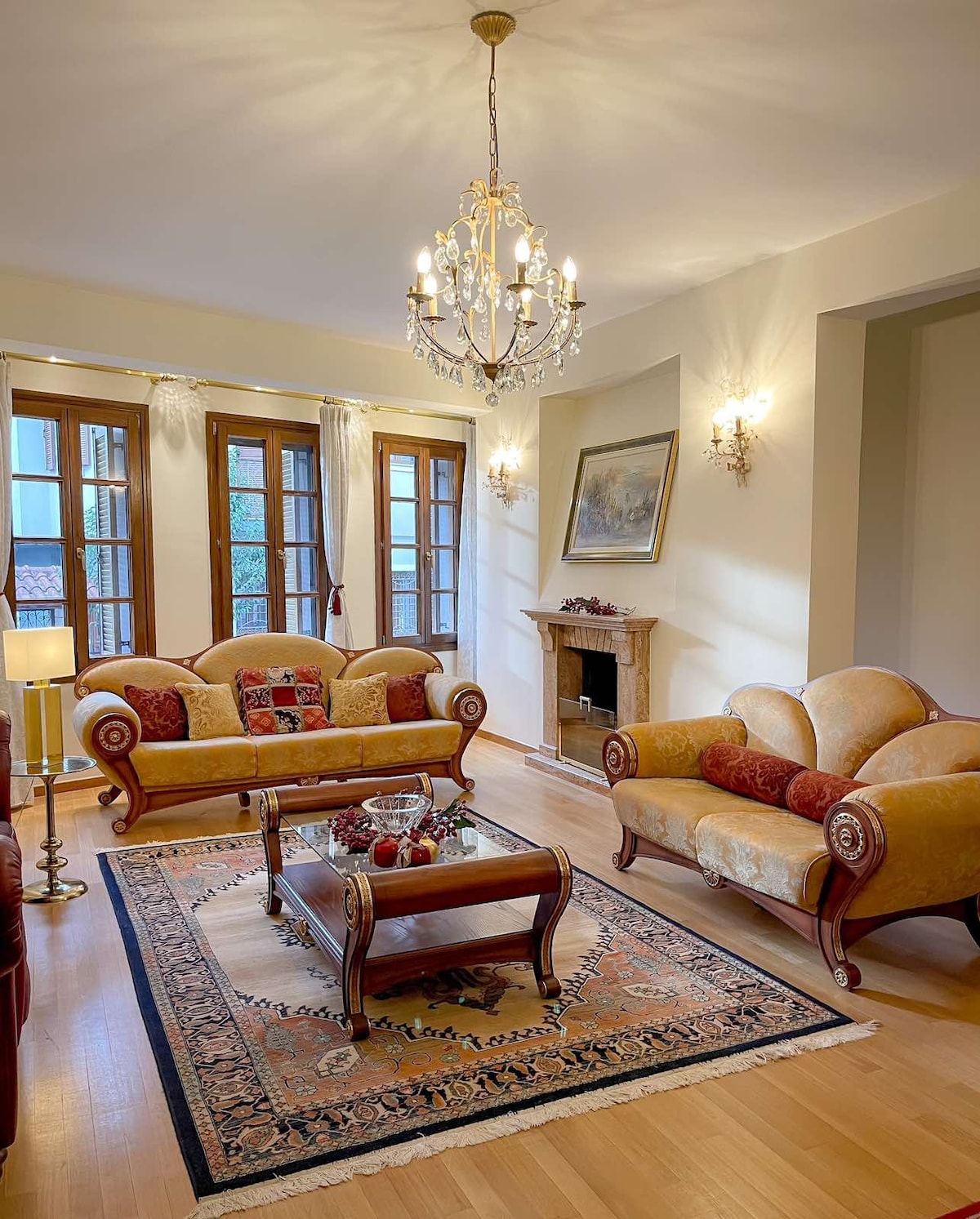 A cozy living room features two elegantly styled sofas with decorative cushions and a central coffee table. Natural light filters through several windows. A chandelier hangs above, and a classic fireplace adds warmth. Artwork is displayed on the wall, enhancing the refined decor.