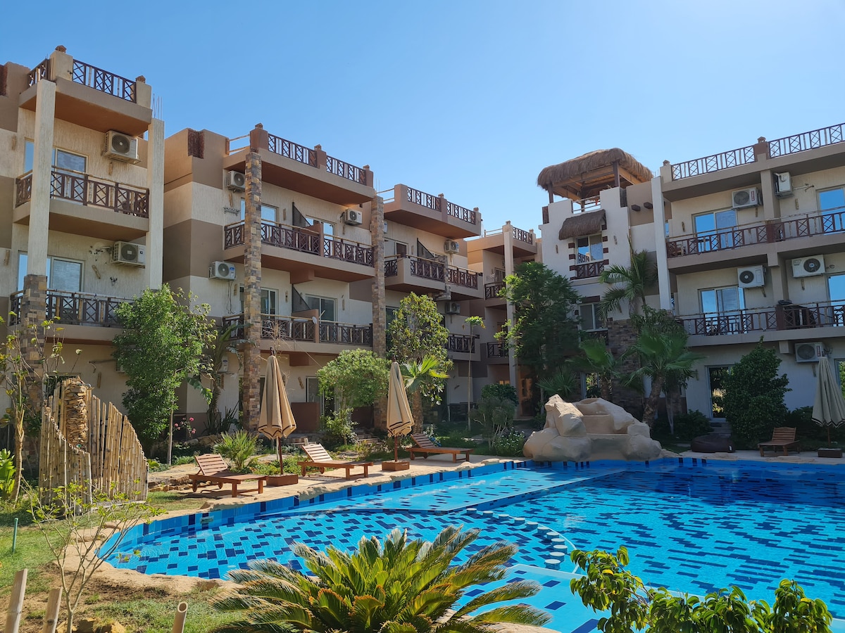 A tranquil courtyard is shown with a large pool surrounded by lush greenery and lounge chairs. The three-story building features balconies and is accented by palms and other greenery, under a clear blue sky.