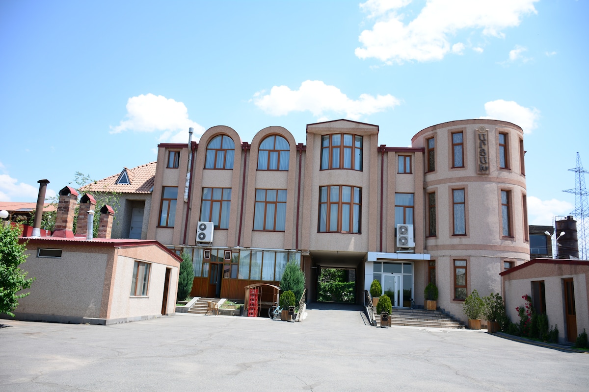 The exterior of a multi-story building is presented, characterized by large arched windows and a distinct round section. A spacious driveway leads to the entrance, bordered by manicured greenery. Air conditioning units are visible on the facade, complemented by subtle architectural details.
