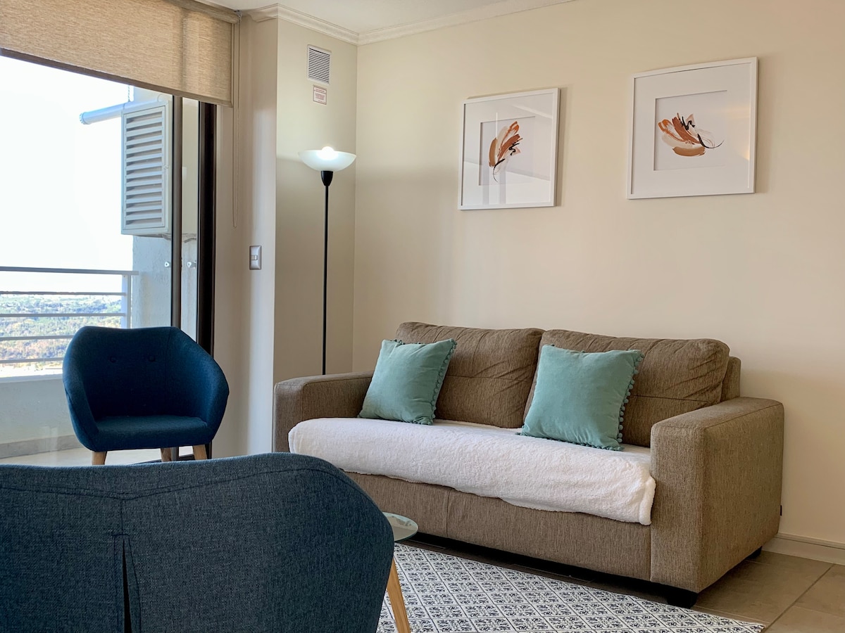 A light-filled living area is presented, featuring a comfortable sofa adorned with two pillows. An accent chair complements the space, while wall art adds a touch of elegance. A lamp provides soft illumination, and large windows offer views of the surrounding area.