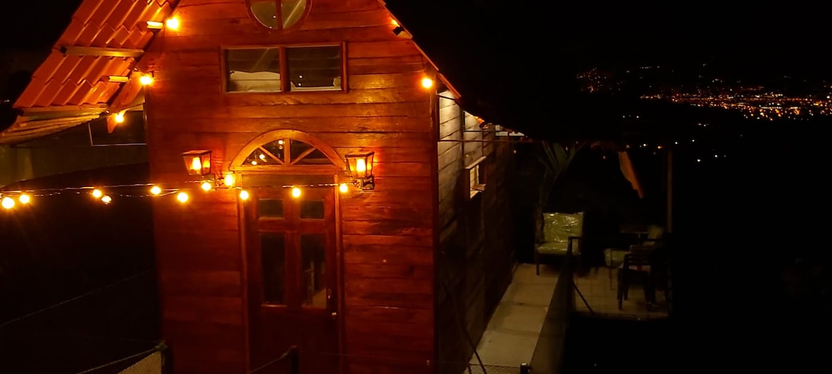 The exterior of a wooden cabin is softly illuminated by warm lights at night, highlighting rustic features and windows. A small seating area with chairs can be seen along the side, set against a backdrop of distant city lights in the valley.