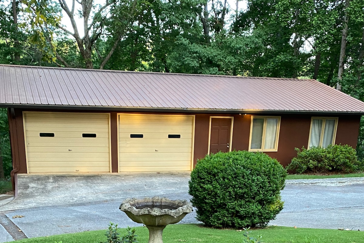 A single-story building features a brown exterior with a metal roof. Two garage doors are situated at the front, alongside a small door. A window is located next to the entrance. Lush greenery surrounds the property, contributing to its natural setting.