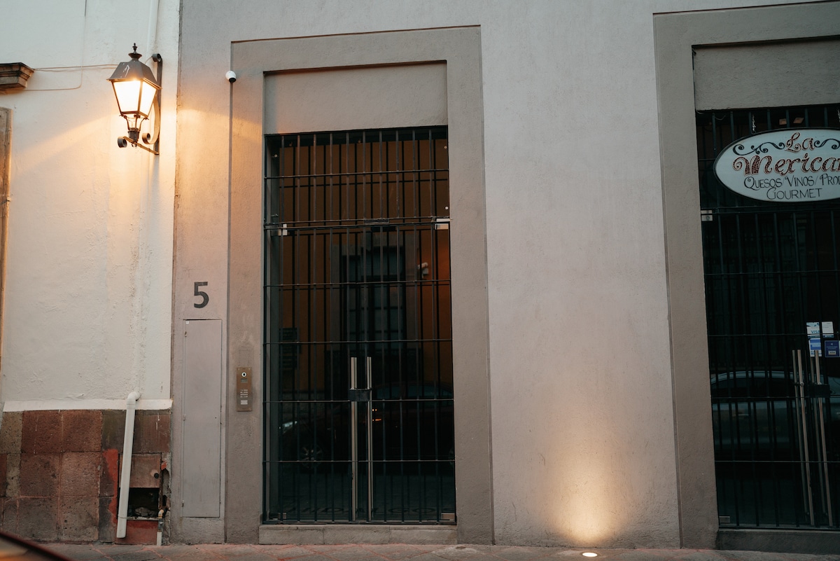 The entrance of the building is marked by a large secure gate framed by a simple light fixture. A small intercom panel is located on the left side of the door, and a decorative sign is affixed beside the entrance, indicating the property name.