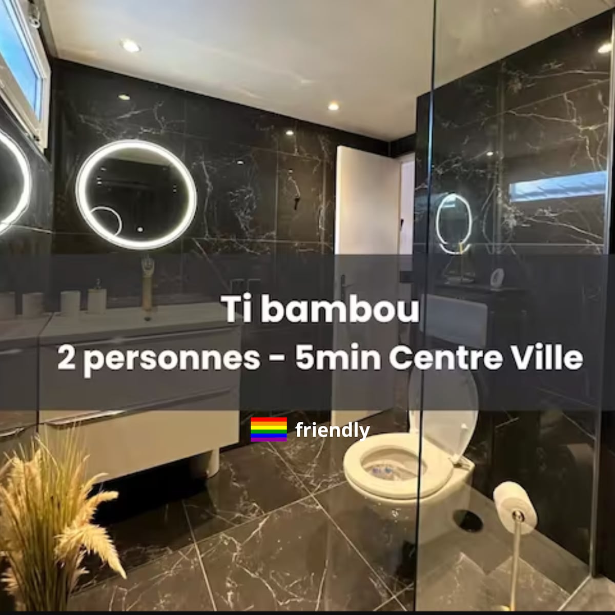 A modern bathroom is presented with sleek black marble tiles and glossy finishes. A round mirror with integrated lighting is mounted above a stylish sink. A minimalist toilet and a glass shower enclosure provide a spacious feel, enhanced by a simple plant arrangement.