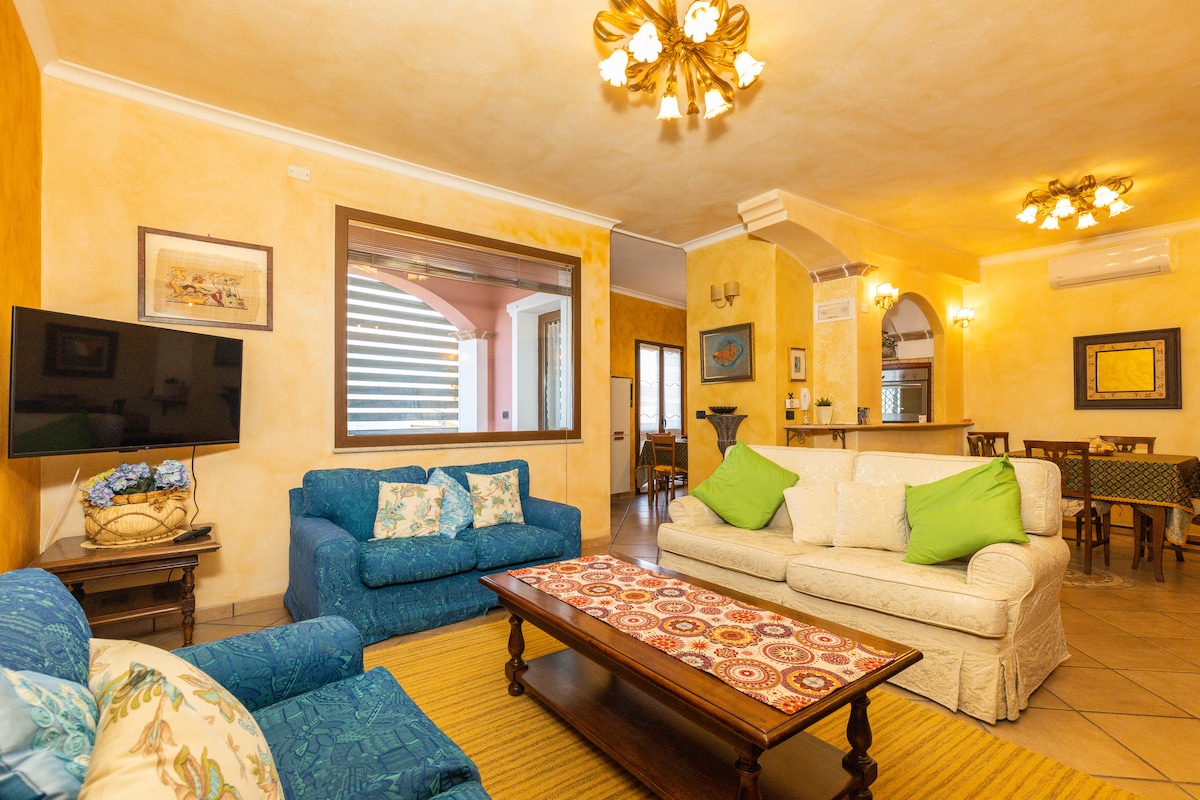 An inviting living space is presented, featuring two blue sofas and a cream-colored couch adorned with green cushions. A decorative coffee table sits in the center, surrounded by warm yellow walls. A large mirror reflects light, while a TV is mounted on the wall opposite the seating area.