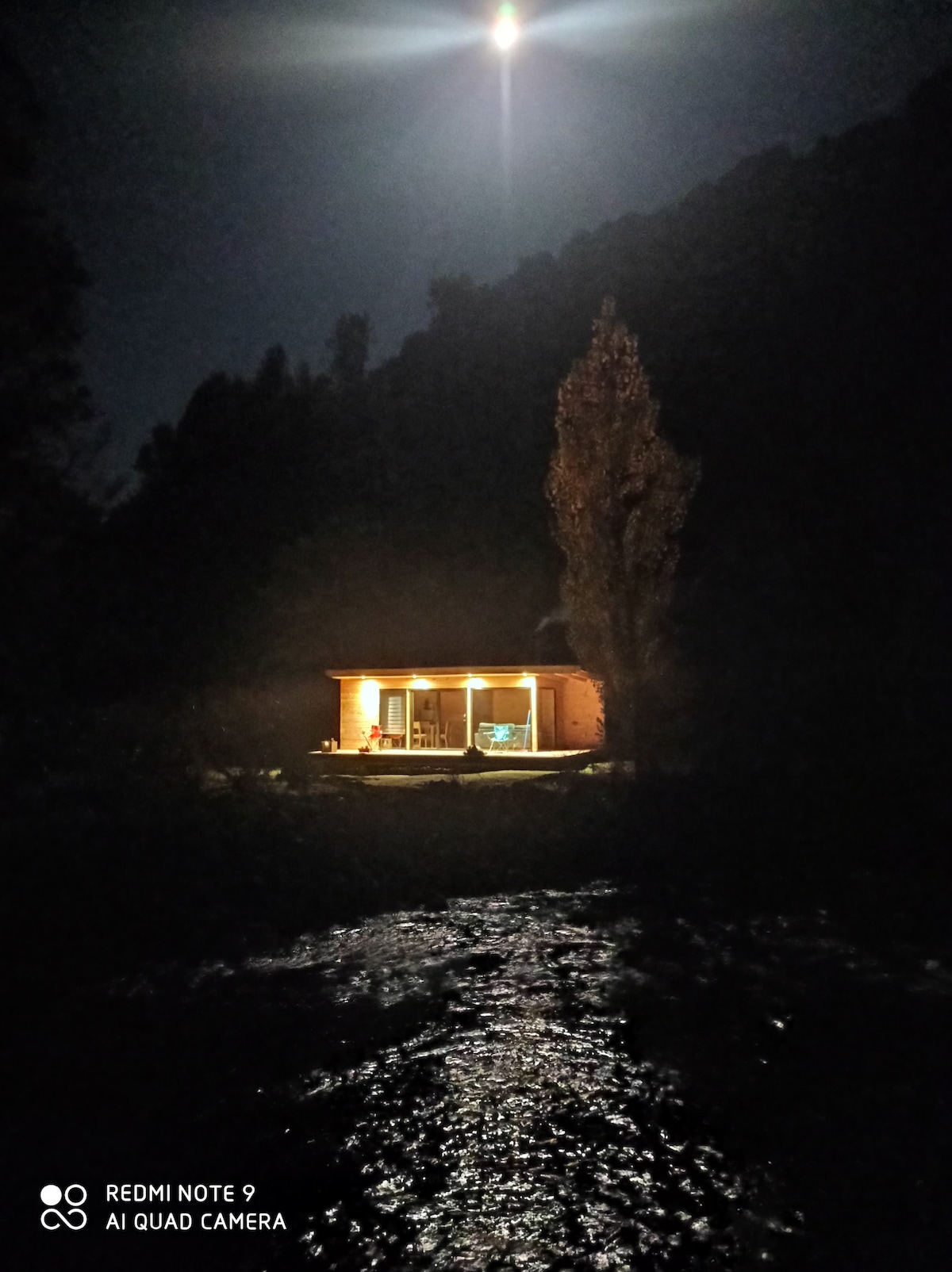 A cabin is softly illuminated by outdoor lights against a dark backdrop of mountains under a night sky. A river flows in the foreground, reflecting the moonlight, while shadows create a serene atmosphere.