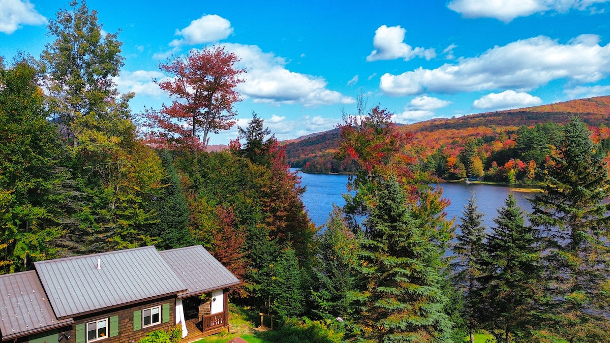 The serene exterior of the studio is framed by vibrant autumn foliage, overlooking the calm waters of Lake Eden. A clear blue sky with scattered clouds enhances the tranquil atmosphere of this rural retreat.