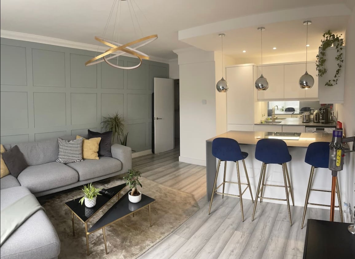 A modern living space showcases a gray sectional sofa adorned with cushions, positioned on a light-colored area rug. An island with barstools is present, complemented by pendant lighting. A green wall adds a touch of nature, and a door leads to another room.