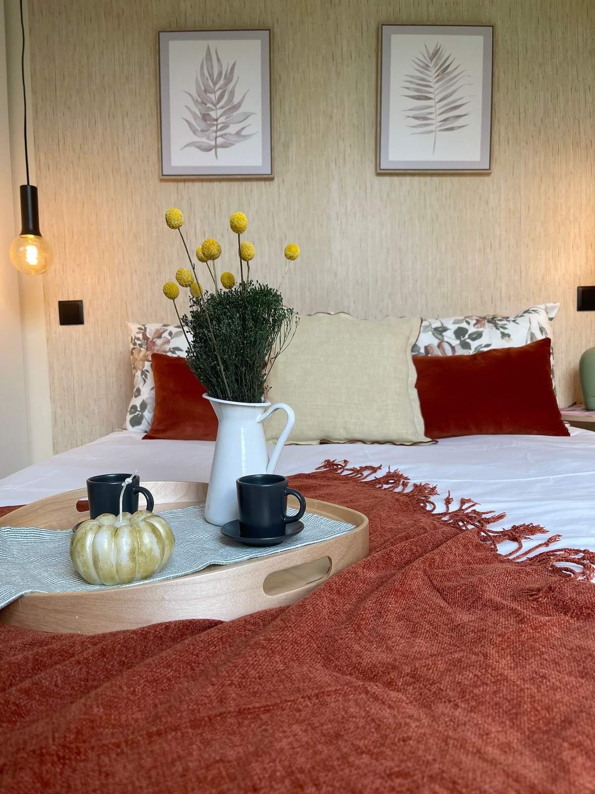 A welcoming sleeping area features a bed adorned with a rust-colored throw and decorative pillows. A wooden tray holds two black mugs, a small pumpkin, and a vase with yellow flowers, complementing two framed botanical prints on the wall.