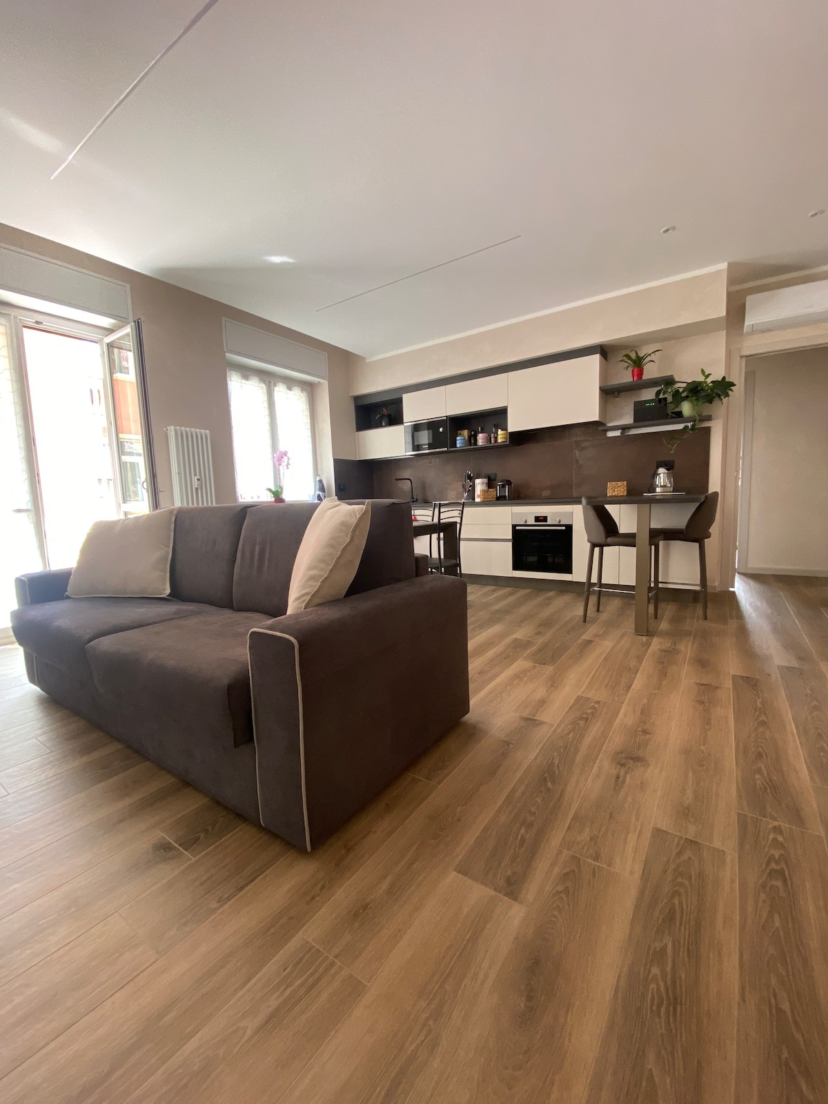 A modern living area features a comfortable brown sofa paired with light cushions. In the background, a well-equipped kitchen is visible, complete with sleek cabinetry and a dining table. Natural light enters through large windows, enhancing the open layout.