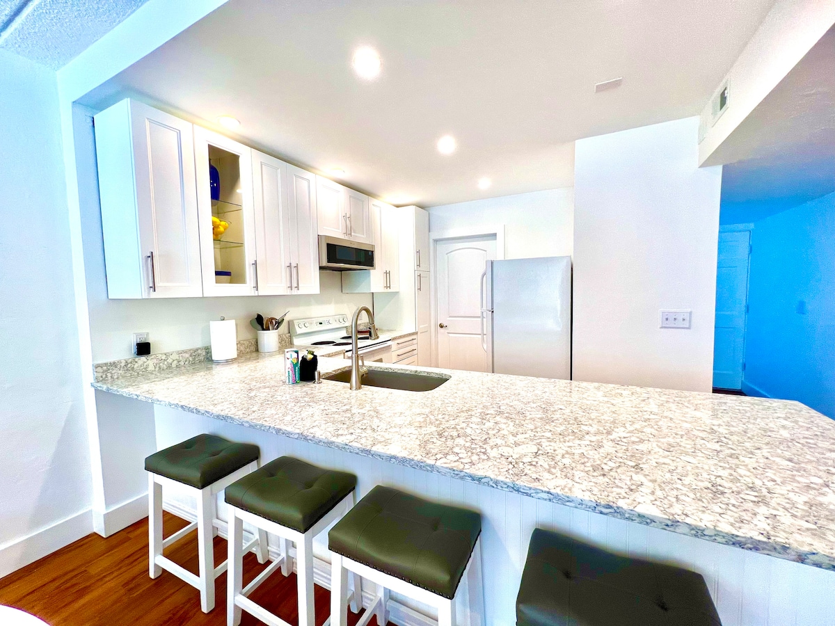 A modern kitchen features a spacious granite countertop with seating for four on padded stools. Sleek white cabinets, stainless steel appliances, and a double sink create a functional and inviting cooking space. Bright overhead lights illuminate the area, enhancing the clean aesthetic.