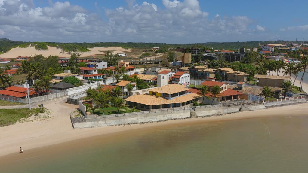 Aerial view of the property showcases a spacious beachside home surrounded by palm trees and sandy shores. Dunes rise in the background, while the shoreline gently curves along clear waters, creating a serene coastal environment.