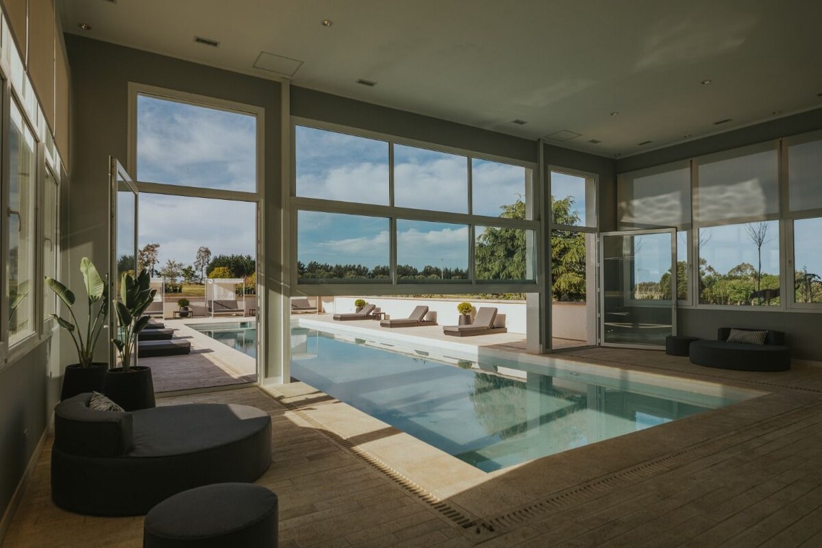 An indoor pool is surrounded by large windows, offering views of the outdoor landscape. Comfortable seating is arranged along the pool area, promoting relaxation. The light-filled space features a neutral color palette and minimalistic design, creating a serene atmosphere.