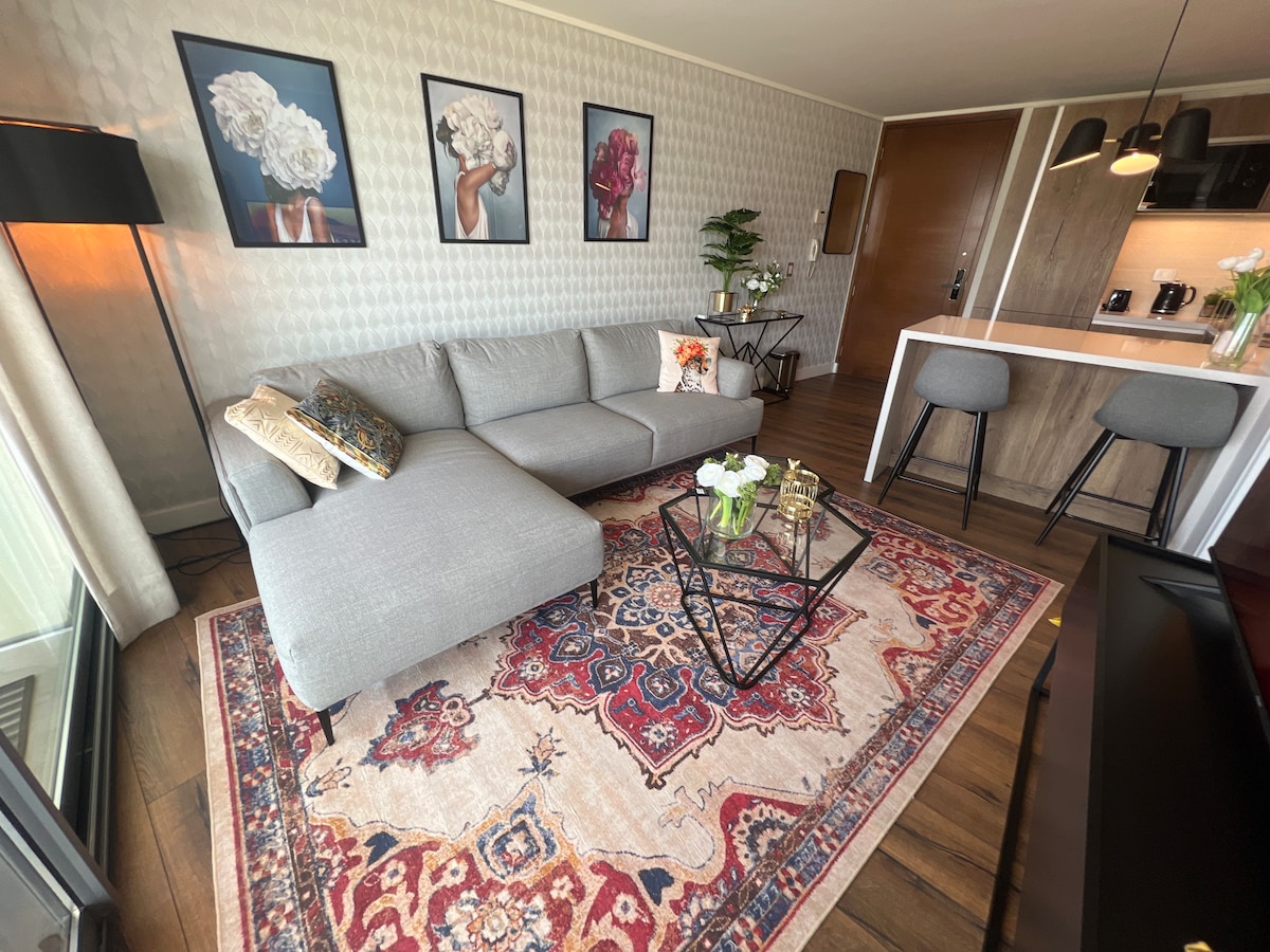 A modern living area features a gray sectional sofa arranged on a decorative area rug. Framed floral artwork adorns the walls, while a glass coffee table with decorative items is placed in front. A small bar area with stools is visible in the background.