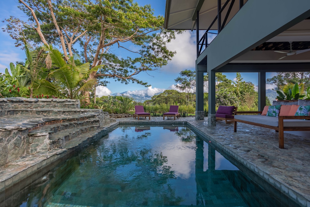 A tranquil saltwater pool is framed by natural stone, with comfortable seating options nearby. Lush greenery and tall trees provide scenic views, while the tranquility of the outdoor space is accentuated by clear skies and gentle reflections on the water's surface.