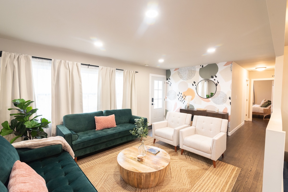 A welcoming living room is presented with a green sectional sofa and two white armchairs facing a wooden coffee table. Natural light enters through large windows, and soft, light-colored curtains frame the space. A colorful wall mural adds a touch of creativity to the atmosphere.