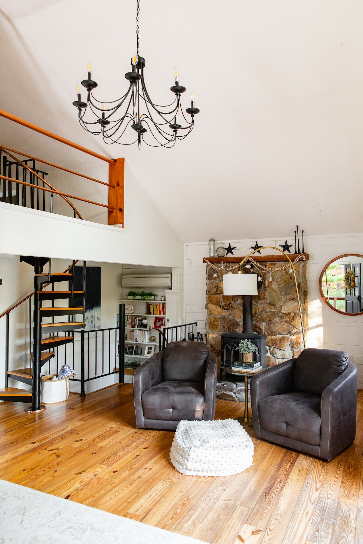 A spacious living area features wooden flooring and a spiral staircase leading to the upper level. Two plush armchairs are positioned beside a stone fireplace, with a cozy ottoman nearby. Natural light fills the room, accentuated by a black chandelier overhead.