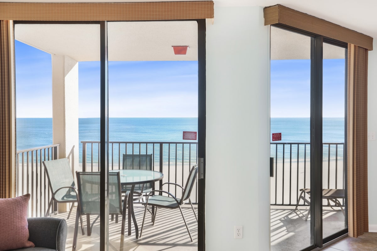 Sliding glass doors open to a private balcony, revealing panoramic ocean views. A round dining table with four chairs is positioned on the balcony, providing a perfect spot to enjoy meals while overlooking the white sandy beach and calm waters of the Gulf.