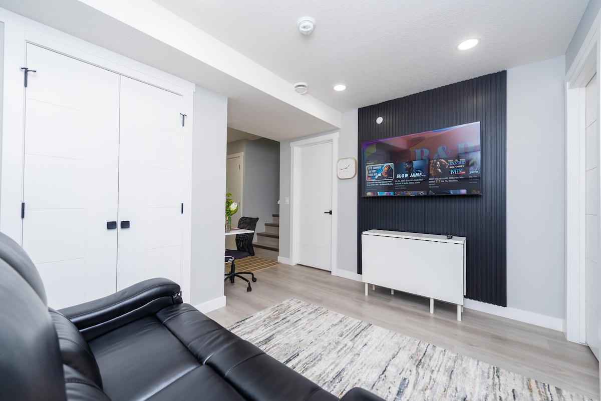 A modern living space features a comfortable leather sofa positioned in front of a flat-screen television mounted on a textured black wall. A decorative area rug adds warmth to the light-colored flooring, and a compact workspace is visible in the background.