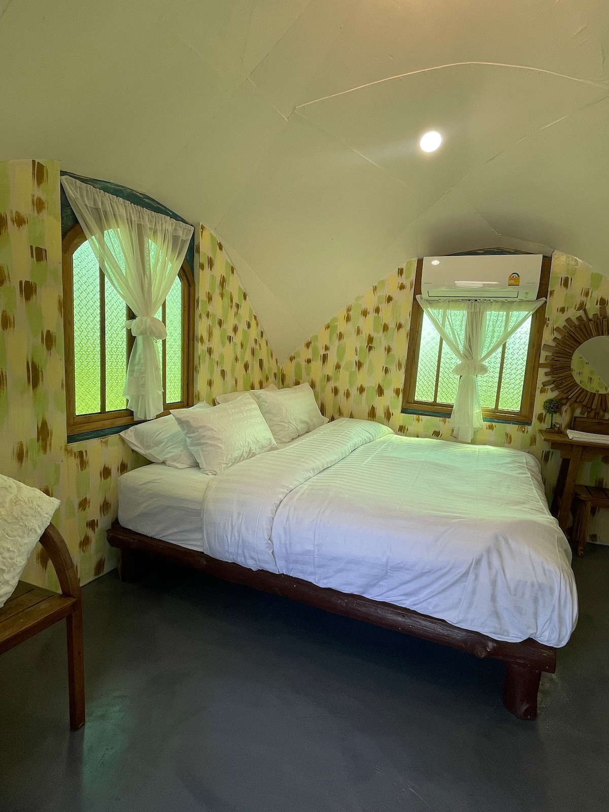 A cozy guest room features a spacious bed with crisp white linens, complemented by patterned walls. Two windows allow natural light to fill the space, with sheer curtains providing a soft touch. An air conditioning unit and a wooden vanity are also visible.