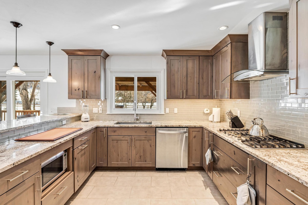 An open plan kitchen features modern wooden cabinetry paired with a light-colored granite countertop. Stainless steel appliances, including a stove and dishwasher, are visible. Natural light streams through the window, illuminating the space. Pendant lights hang above the central island, enhancing functionality.
