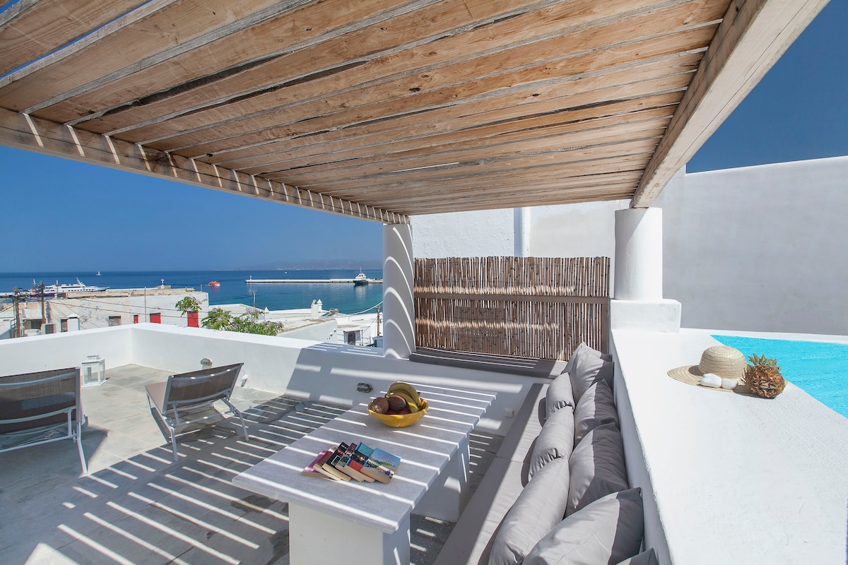 An outdoor terrace is shown, featuring a shaded seating area with comfortable cushions and a table set with decorative items. A panoramic view of the sea and nearby waterfront is visible, along with a pool edge reflecting the sky. Sun loungers are positioned beside the seating area.