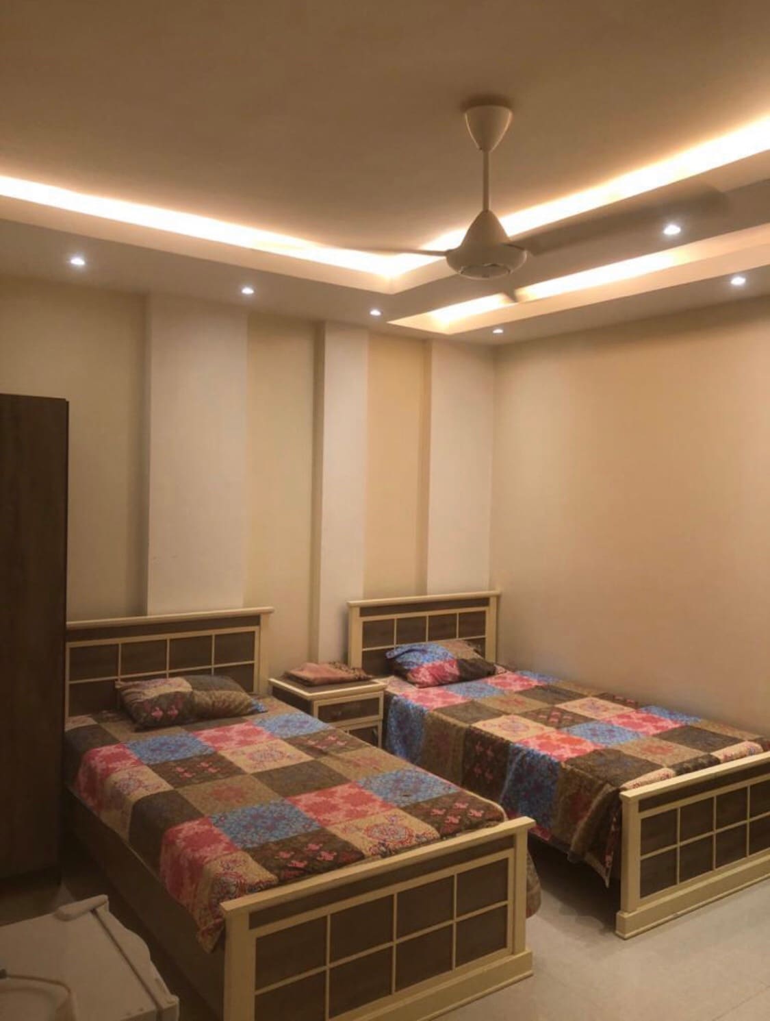 A calm bedroom features two spacious beds with colorful patchwork blankets. Soft lighting from the ceiling highlights the light-colored walls, creating a soothing atmosphere. A bedside table is positioned next to one bed, while a wardrobe is visible in the corner.