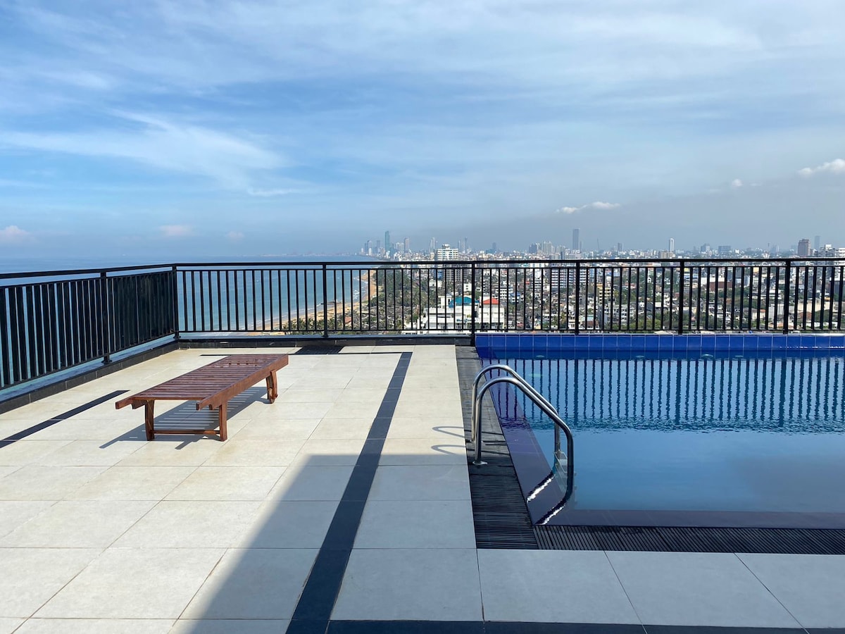 An outdoor pool area features a clean swimming pool with a ladder for easy access. A sun lounger is positioned on the tiled deck, offering views of the surrounding skyline and ocean horizon. The area is enclosed by a black railing.