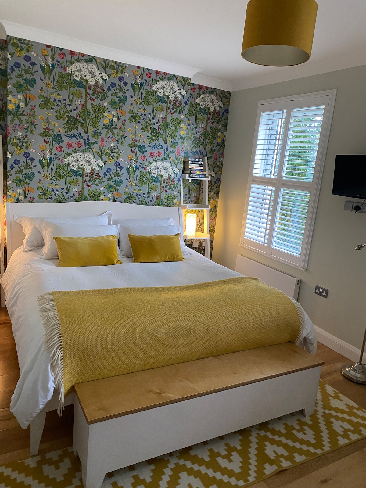 A bright room features a spacious bed dressed in crisp white linens and accented with yellow cushions. A patterned wallpaper of floral design enhances the ambiance. Natural light enters through the shutters of a large window, while a rug and a bench add functional elements to the space.
