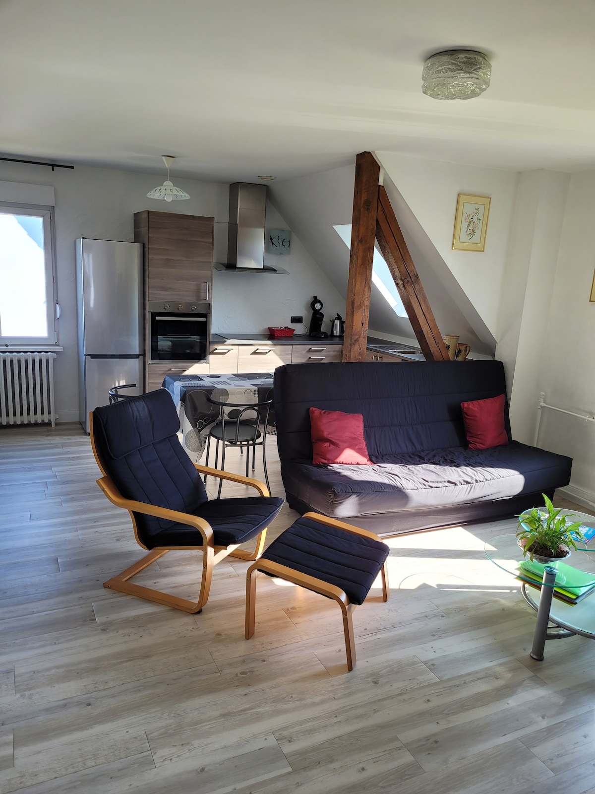An open-plan living area features a dark sofa and a wooden armchair with red cushions. A sleek kitchen is visible in the background, equipped with modern appliances. Natural light fills the space through large windows, highlighting the wooden beams and light-colored flooring.