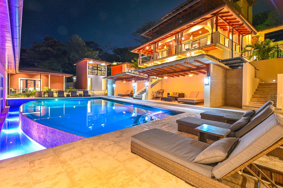A beautifully illuminated pool area is featured, showcasing an infinity pool with lighting that highlights its contours. Cozy lounge chairs are positioned by the water. Surrounding structures display a mix of vibrant colors, with outdoor seating available for relaxation and enjoyment.