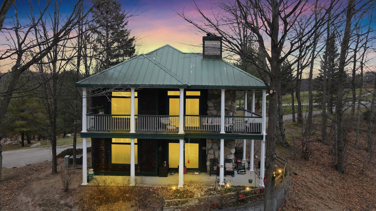 A charming two-story condo features a green metal roof and stone accents. Large windows emit a warm glow, highlighting the inviting porch. Surrounding trees frame the structure, which is set in a serene landscape with a hint of twilight in the sky.