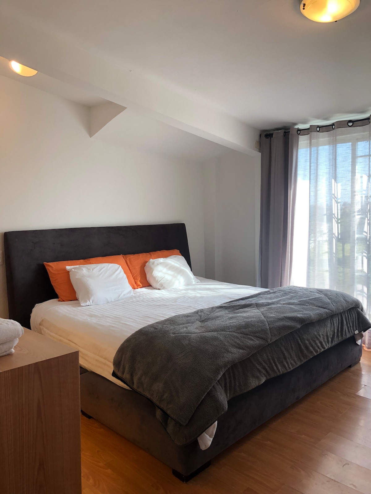 A cozy bedroom features a spacious bed with contrasting orange and white pillows. Natural light enters through large windows, which are dressed in light curtains, creating a bright and airy environment. A simple wooden nightstand adds functionality to the space.