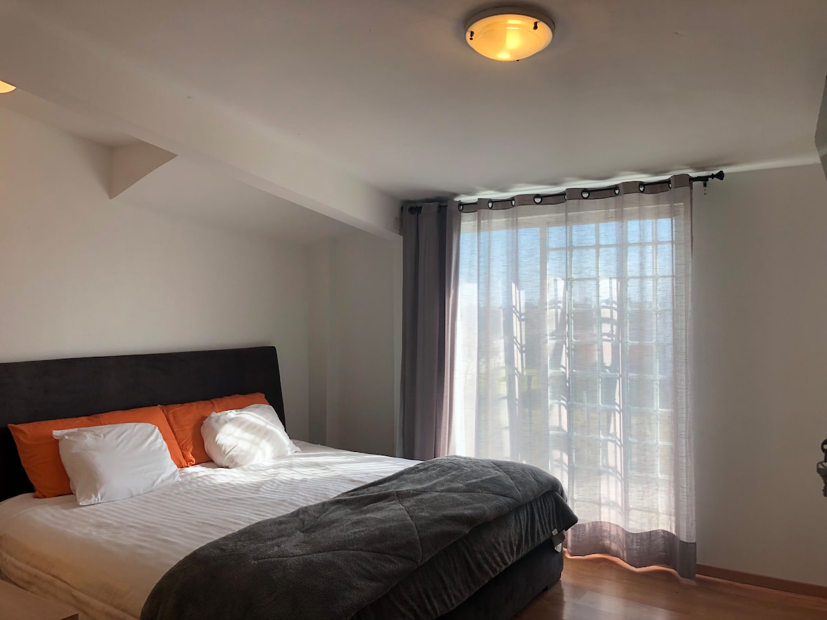 A spacious sleeping area features a large bed with white linens and orange accent pillows. Natural light filters through sheer curtains, illuminating the room. A soft gray blanket is draped over the bed, complementing the wood flooring and neutral walls.