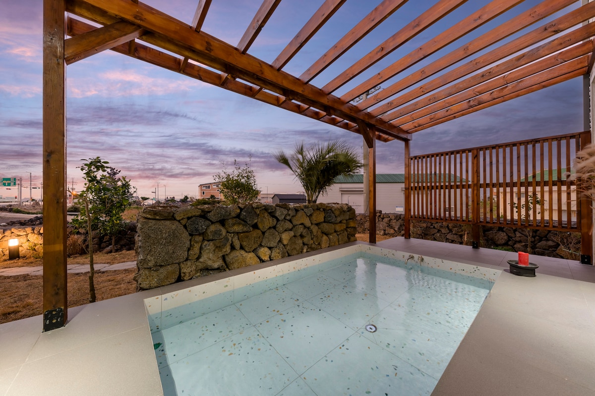 An outdoor hot tub is situated beneath a wooden pergola, surrounded by stone walls. The space features a smooth, light-colored tiled deck and is framed by a landscape of trees and grass. A serene sky with soft hues of purple and pink is visible above.