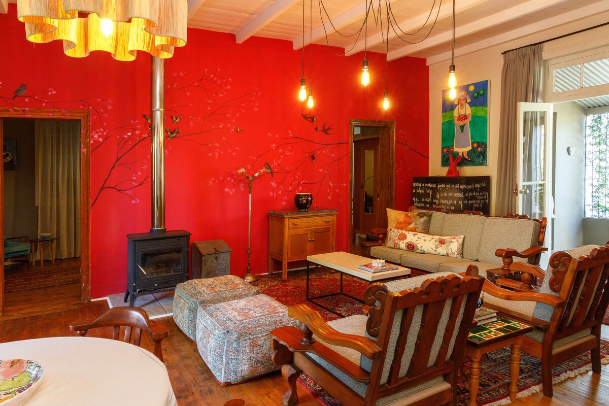 A vibrant living area features a striking red accent wall adorned with decorative blossoms. Comfortable seating is arranged around a central coffee table, complemented by various textures in the furnishings. A wood-burning stove provides warmth, while soft light is emitted from overhead pendant lights.