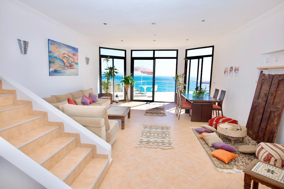 The spacious living area showcases large glass doors that open onto the terrace with ocean views. A light-colored sofa is arranged with colorful cushions, complemented by a table and dining chairs. Decorative rugs and plants add a touch of warmth to the environment.