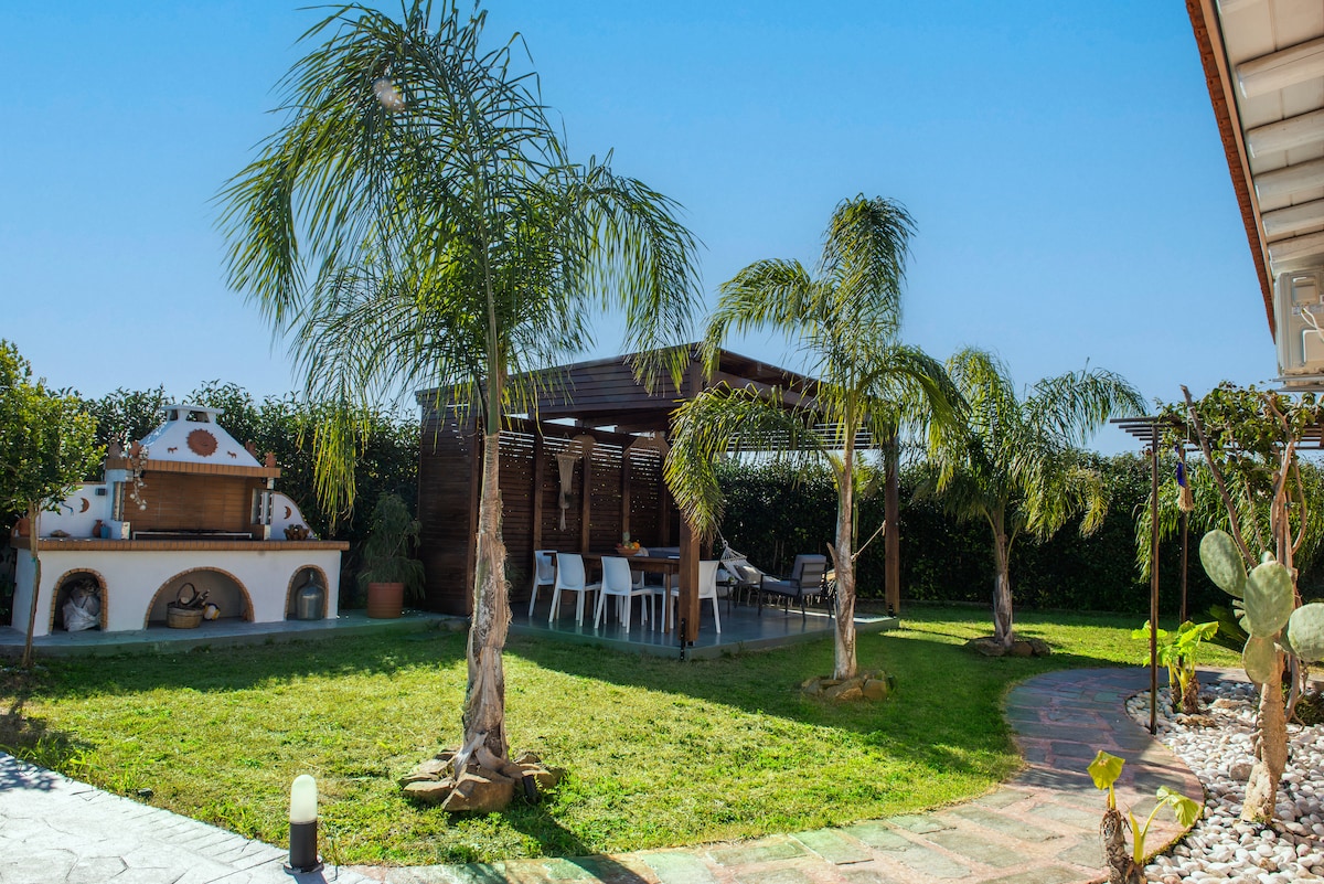 A well-maintained garden area features lush green grass and palm trees, providing shade. A wooden pergola is visible, adorned with tables and chairs, ideal for outdoor dining. A barbecue station complements the space, surrounded by assorted plants and pathways.