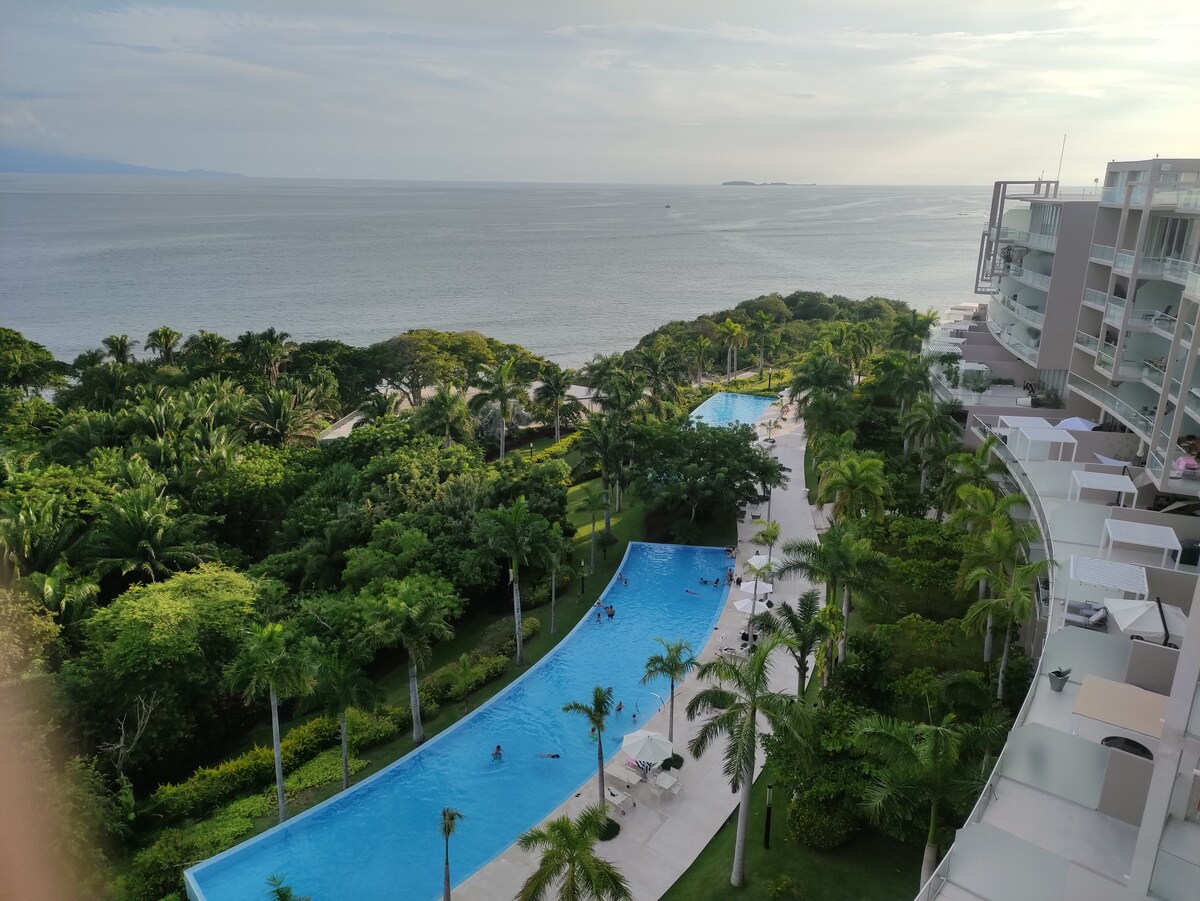 The image captures a serene view overlooking lush greenery and a winding blue pool that leads to the ocean. Palm trees line the area, creating a tropical ambiance. The horizon features water extending into the distance under a partly cloudy sky.