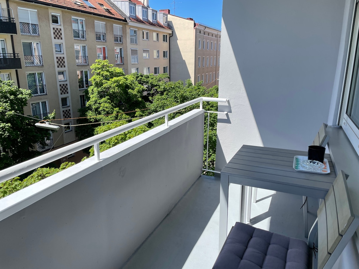 A balcony with a minimalist design features a small table and a chair set. Lush greenery from nearby trees is visible, along with views of surrounding buildings. Natural light illuminates the space, creating a bright and airy atmosphere.
