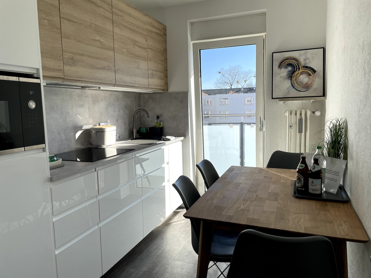 A modern kitchen space features sleek white cabinetry and wooden accents. Natural light enters from a balcony door, illuminating a wooden dining table with black chairs. Kitchen appliances such as a microwave and stovetop are visible, alongside essentials for cooking and dining.