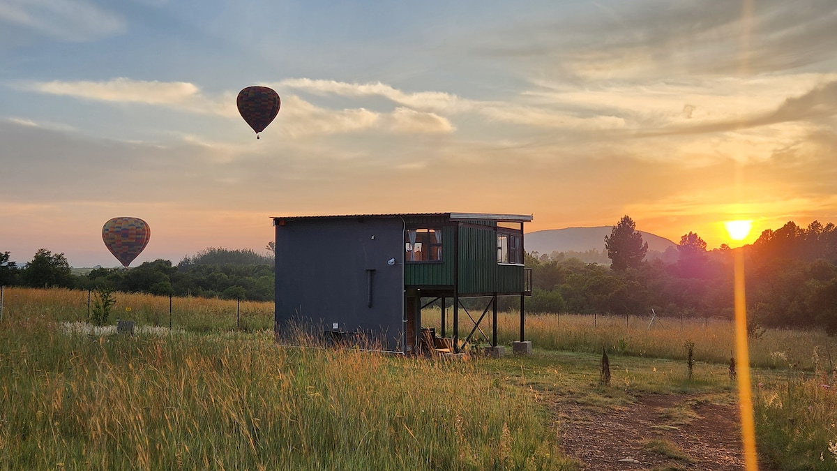 A raised unit stands amidst tall grass, framed by a warm sunrise. Two colorful hot air balloons float in the sky, while the backdrop features rolling hills and a serene landscape, creating a peaceful and open atmosphere.
