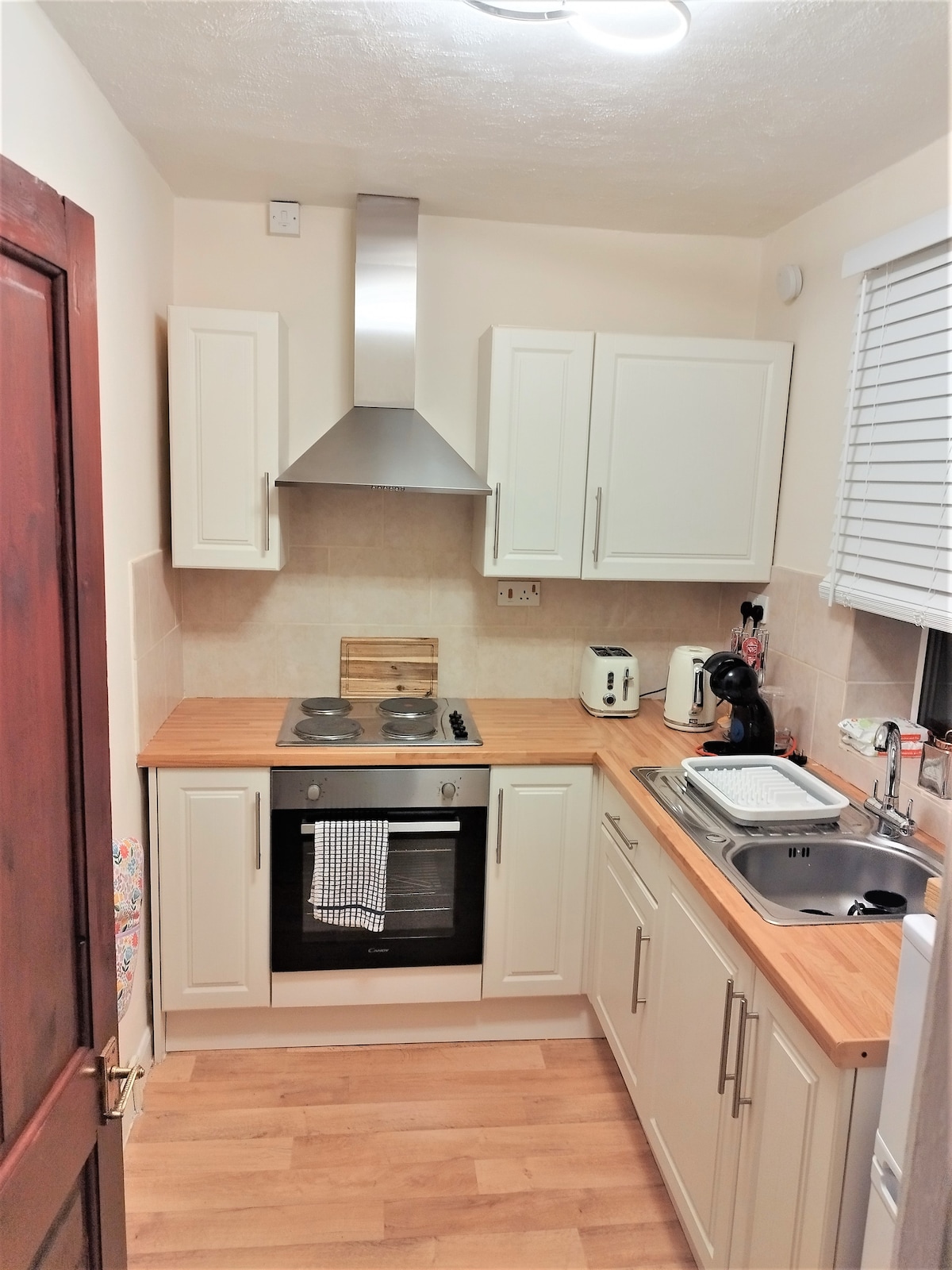A fully equipped kitchen features modern cabinetry with a warm wood countertop. Stainless steel appliances include an oven and a kettle, while natural light filters through a shaded window. Dishware and kitchen utensils are neatly arranged for convenience.