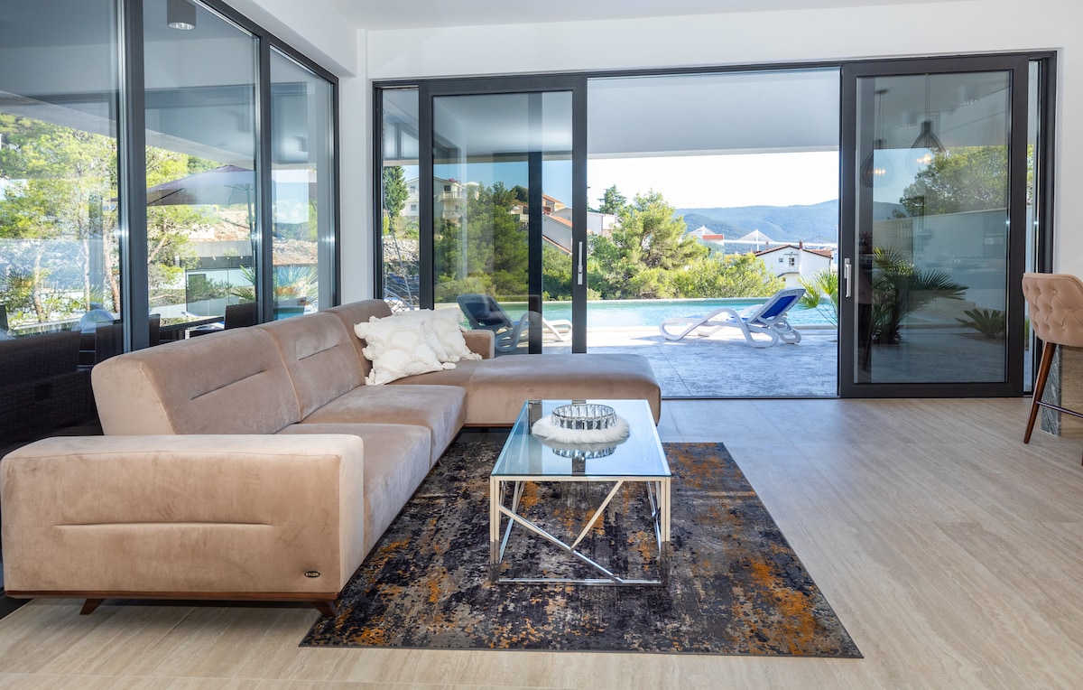 A spacious living area features a modern beige sofa and a glass coffee table. Large sliding doors provide a view of the pool and surrounding greenery, creating a seamless connection with the outdoors. A decorative rug adds warmth to the light-colored floor.