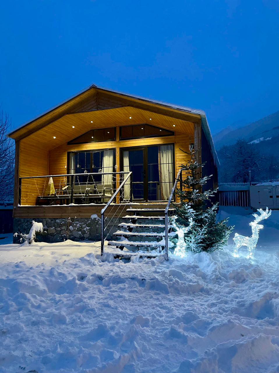 A modern villa is showcased, featuring a wooden exterior and large glass doors. Snow blankets the surrounding area, while a festive reindeer decoration and a small tree are illuminated in soft white light, creating a serene winter ambiance.