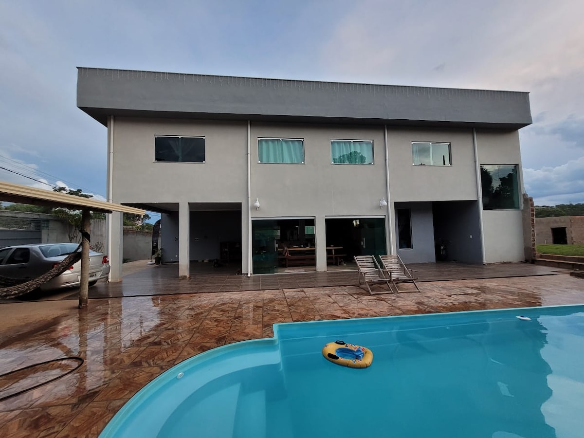 A modern two-story building features expansive glass windows, allowing natural light to fill the interior. A well-maintained pool with a floating toy is visible in the foreground, reflecting the clear sky. A spacious patio is accessible from the large glass doors.