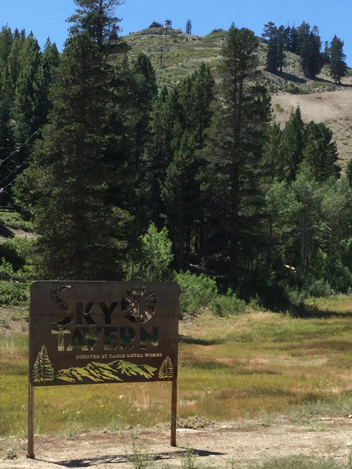 A weathered sign marks the entrance to Sky Tavern, surrounded by lush green grass and scattered trees. Towering mountains can be seen in the background, providing a sense of the natural landscape. Bright sunlight highlights the surrounding area, creating a welcoming environment.