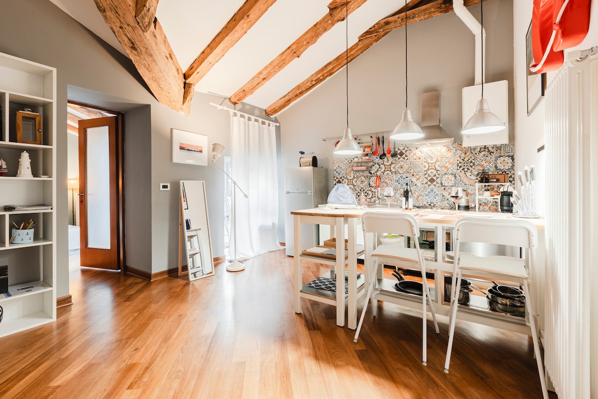 A well-equipped kitchen area features a wooden dining table surrounded by white chairs. Two pendant lights hang above, illuminating the colorful backsplash tiles. Natural light filters through a nearby curtain, highlighting the warm wood floors and exposed wooden beams above.