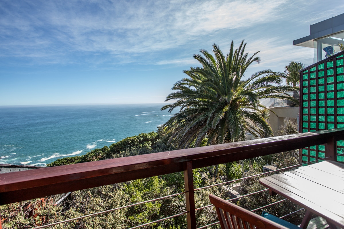A timber deck offers a sweeping view of the ocean, framed by lush greenery and palm trees. The clear blue sky complements the vibrant colors of the landscape, creating a serene outdoor space ideal for relaxation.